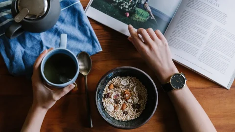 Overhead view of a person's hands with one holding a mug of tea and the other resting on a book beside a bowl of healthy cereal with nuts and seeds on a wooden table Healthy Eating habits