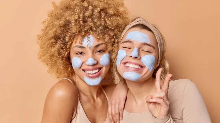 Two women with diverse skin tones applying blue face masks and smiling, showcasing the benefits of skincare routines including Vitamin C for radiant and youthful skin.